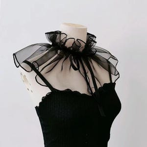 Pode incluir: Uma gola preta com babados de tecido transparente, com um laço de fita, apresentada em um manequim. A gola é feita de um tecido delicado e fluido, e o design é em camadas para maior volume. A gola é combinada com um top preto.