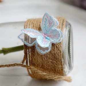 May include: A light blue butterfly-shaped lace applique with a pink and blue design. The applique is attached to a brown twine-wrapped jar.