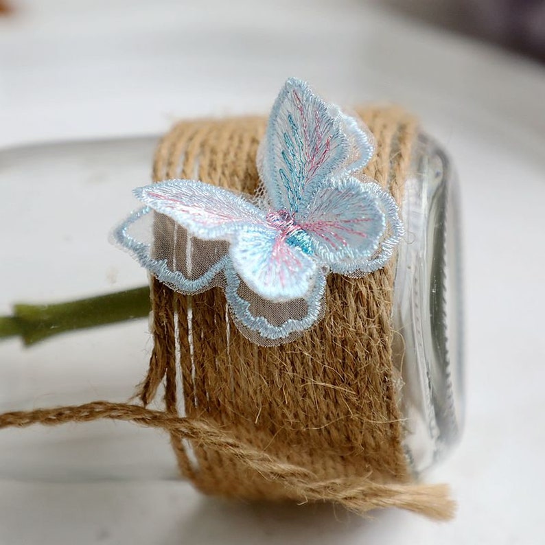 May include: A light blue butterfly-shaped lace applique with a pink and blue design. The applique is attached to a brown twine-wrapped jar.