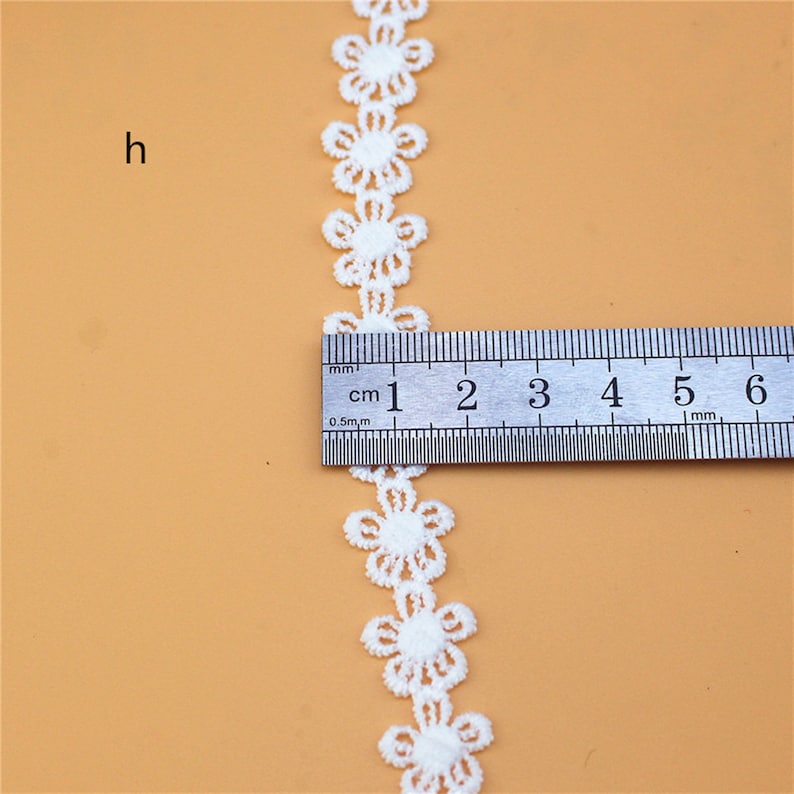 May include: White lace trim with a repeating flower design. The trim is measured with a ruler, showing it is about 1 inch wide.