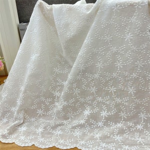 Puede incluir: Una tela de algod&oacute;n blanca con un intrincado patr&oacute;n floral bordado en toda la superficie. El tejido muestra delicadas flores y enredaderas cosidas, terminado con un borde festoneado a lo largo de la parte inferior del material.