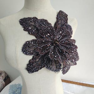 Può includere: Una grande applicazione floreale di colore viola scuro realizzata con paillettes e perline. L'applicazione è perfetta per aggiungere un tocco di brillantezza ed eleganza a qualsiasi capo di abbigliamento.