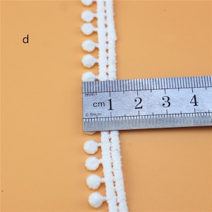 May include: White cotton trim with small pom-poms. The trim is measured with a ruler showing it is 1 cm wide.