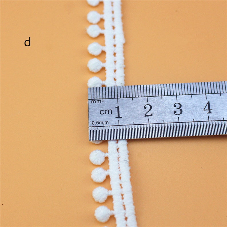 May include: White cotton trim with small pom-poms. The trim is measured with a ruler showing it is 1 cm wide.