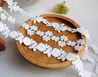 Op de afbeelding: Witte bloemen kanten boord met een herhalend patroon van rozen en kleine bloemen. Het kant wordt getoond op een houten bord, wat het delicate ontwerp benadrukt. Ideaal voor knutselen, naaien of het toevoegen van een decoratief tintje.