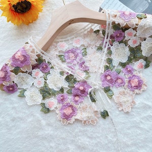 May include: White lace collar with a floral design featuring purple, pink, and green embroidery. The collar has a tie closure.