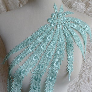 Może przedstawiać: Aplikacja z koronki w jasnoniebieskim kolorze z wzorem piór pawia. Aplikacja ma duży, skomplikowany wzór kwiatowy u góry i kaskadowe pióra z ozdobami w kształcie łez.