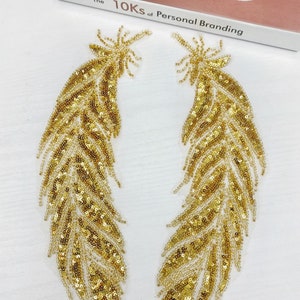 May include: Two gold sequined feather appliques. Each applique features a detailed design with small, shiny sequins and beads, creating a textured and shimmering effect. The appliques are likely for embellishing clothing or accessories.