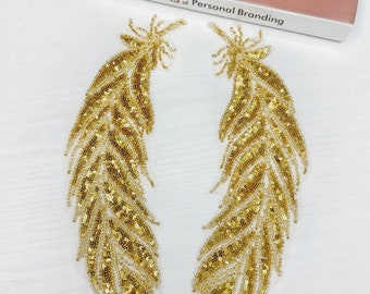 May include: Two gold sequined feather appliques. Each applique features a detailed design with small, shiny sequins and beads, creating a textured and shimmering effect. The appliques are likely for embellishing clothing or accessories.