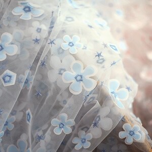 May include: White sheer fabric with a blue floral pattern. The flowers are 3D and have a white outline. The fabric is delicate and has a soft texture.
