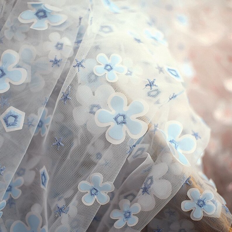 May include: White sheer fabric with a blue floral pattern. The flowers are 3D and have a white outline. The fabric is delicate and has a soft texture.
