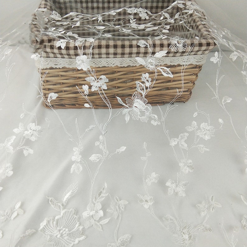 K&ouml;nnte beinhalten: Wei&szlig;er Blumenspitzenstoff mit einem zarten Muster. Die Spitze ist &uuml;ber einen Weidenkorb mit einer braunen und wei&szlig;en karierten Auskleidung gelegt.