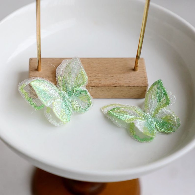May include: Two green and white embroidered butterfly appliques on a white surface.