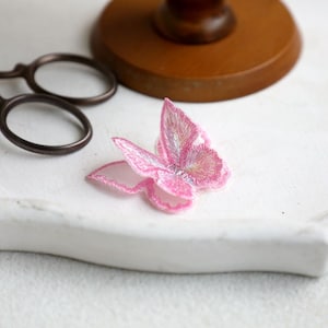 May include: A delicate pink butterfly-shaped lace applique with a shimmery finish. This embellishment is perfect for adding a touch of whimsy to crafts and sewing projects.
