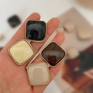 Peut inclure: Une main tenant quatre boucles d'oreilles carrées de couleurs variées : noir, crème, marron et beige. Chaque boucle d'oreille a un cadre en métal doré. Les boucles d'oreilles ont un aspect marbré ou brillant, suggérant de la résine ou de l'émail. Accessoire de mode.