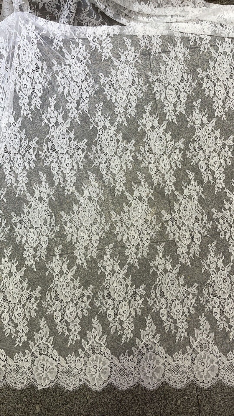 May include: White floral lace fabric with a scalloped edge. The lace has a delicate floral pattern and is perfect for crafting, sewing, and decorating.