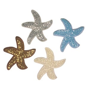 Op de afbeelding: Vier pailletten zeesterren in zilver, blauw, goud en crème. Elke zeester heeft een gestikte omtrek en is gevuld met glinsterende pailletten. De zeesterren zijn gerangschikt op een witte achtergrond.