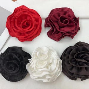 May include: Five fabric rose-shaped accessories in red, burgundy, black, white, and brown. Each rose has layered petals, creating a three-dimensional effect. The accessories are likely for clothing or craft projects.