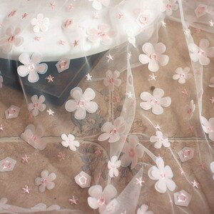 May include: White tulle fabric with pink 3D flower embellishments. The fabric has a delicate, feminine look and would be perfect for crafting or sewing projects.