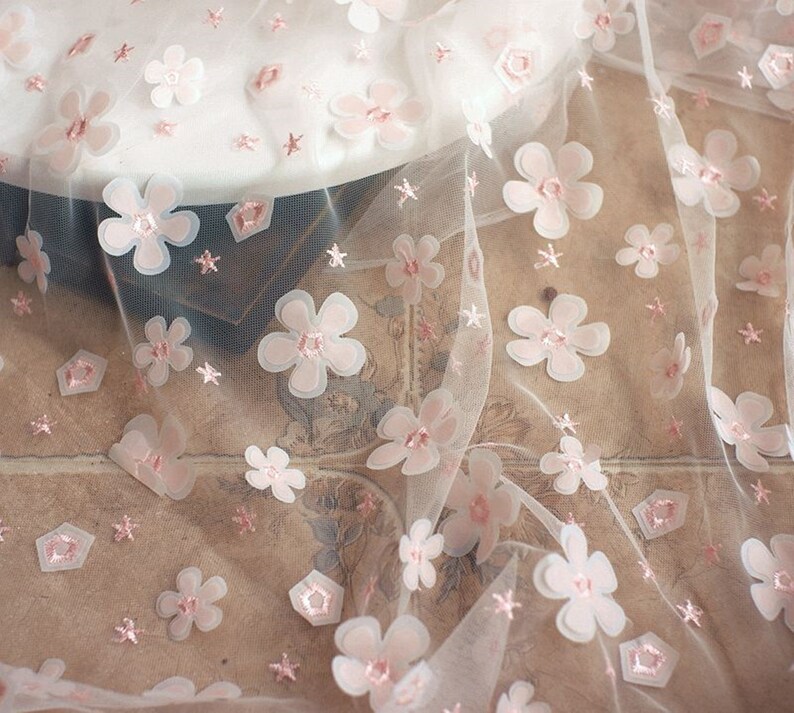 May include: White tulle fabric with pink 3D flower embellishments. The fabric has a delicate, feminine look and would be perfect for crafting or sewing projects.