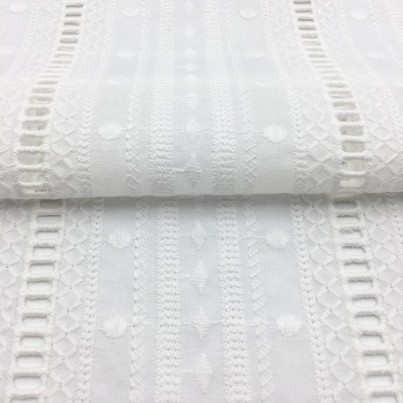 May include: White cotton fabric with an embroidered pattern of geometric shapes and small circles. The fabric has a delicate, lacy look.