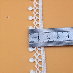 May include: White lace trim with a scalloped edge and small white pom-poms. The trim is measured with a ruler showing 1 cm.