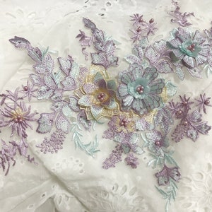 Puede incluir: Una delicada aplicación floral con flores y hojas de color pastel morado, azul y amarillo. La aplicación está adornada con pequeñas cuentas de perlas.