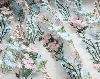May include: Delicate floral embroidered fabric featuring light blue and pink flowers with green stems and leaves. The embroidery is on a sheer white background, creating a soft, airy look. Ideal for crafting or apparel.