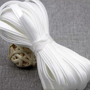 Peut inclure: Une bobine de ruban de biais blanc à double pli en tissu lisse, présentant une largeur constante et une finition nette. Le ruban est soigneusement enroulé en un paquet et fixé par un petit lien en tissu blanc assorti au centre de la bobine.