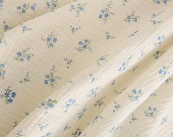 Op de afbeelding: Een gebroken witte stof met een gekreukte textuur en een herhalend patroon van kleine blauwe bloemen met groene stengels en bladeren. Het materiaal is gedrapeerd om plooien en schaduwen op het oppervlak van het textiel te tonen.