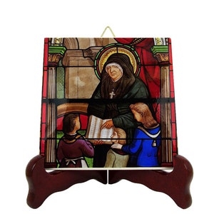May include: A stained glass tile depicting a nun teaching children. The nun is wearing a black habit and a white veil. She is holding a book and pointing to it with her right hand. The children are dressed in colorful clothing. The tile is framed in a gold-colored frame and has a wooden stand.