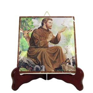 May include: A ceramic tile featuring a depiction of Saint Francis of Assisi surrounded by animals. He is holding a dove in his hand, and there are other animals around him, including a deer, a lamb, and a rabbit. The tile is displayed on a wooden stand.