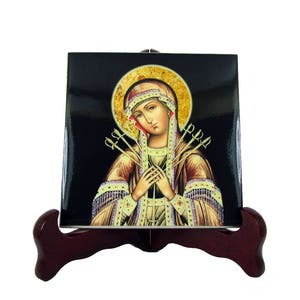 May include: A ceramic tile featuring a religious image of a woman wearing a crown of thorns with seven swords piercing her heart. The tile is displayed on a wooden stand.