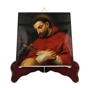 May include: A religious artwork print on a square tile, depicting a man in a red robe holding a cross. The tile is displayed on a dark red wooden easel. The man has a halo and is looking down.