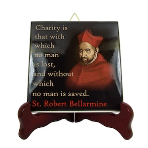 May include: A black and gold ceramic tile with a quote by Saint Robert Bellarmine. The quote reads: "Charity is that with which no man is lost, and without which no man is saved." The tile features a portrait of Saint Robert Bellarmine in a red cardinal's hat and robe.