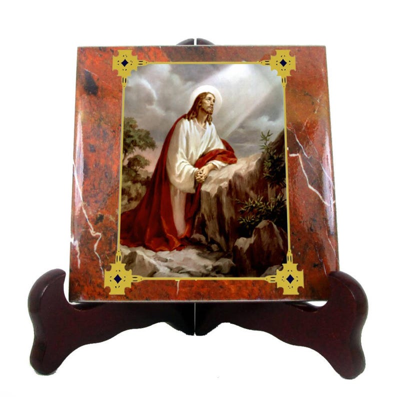 May include: A ceramic tile featuring a picture of Jesus praying in a red robe on a rocky hill. The tile has a gold frame with a decorative design in the corners. The tile is displayed on a wooden stand.