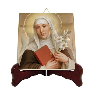 May include: A ceramic tile with a colorful image of a woman in a white habit holding a book and a lily. The tile is on a wooden stand.