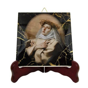 Può includere: Una piastrella di ceramica con sfondo in marmo nero e oro. La piastrella presenta un dipinto di una donna vestita con una tunica bianca e una corona di spine. La piastrella è su un supporto in legno.