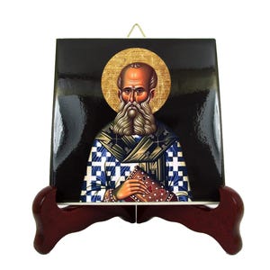Könnte beinhalten: Eine Keramik-Ikone des Heiligen Nikolaus, einer religiösen Figur mit weißem Bart und einem blau-weiß karierten Gewand, die ein Buch hält. Die Ikone befindet sich auf einem dunklen Hintergrund und hat einen goldenen Heiligenschein um den Kopf des Heiligen. Die Ikone wird auf einem Holzständer präsentiert.