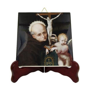 Può includere: Una piastrella di ceramica con un'immagine religiosa di un santo che tiene un libro e un crocifisso. Il santo indossa una veste marrone e ha un'aureola. Un cherubino tiene il libro e guarda il crocifisso. La piastrella è su un supporto di legno.