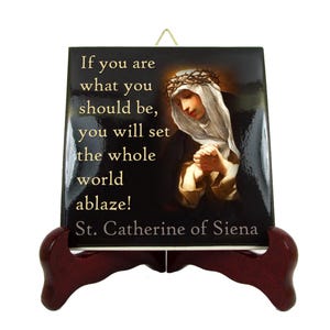 Può includere: Una piastrella di ceramica nera e dorata con una citazione di Santa Caterina da Siena: "Se sei ciò che dovresti essere, darai fuoco al mondo intero!" La piastrella presenta un dipinto di Santa Caterina da Siena con una corona di spine e uno sfondo nero.