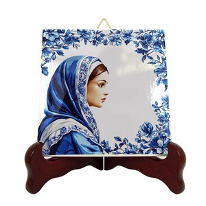 Op de afbeelding: Een keramische tegel met een blauwe en witte bloemrand en een portret van een vrouw met een blauwe hoofddoek. De tegel staat op een houten standaard.