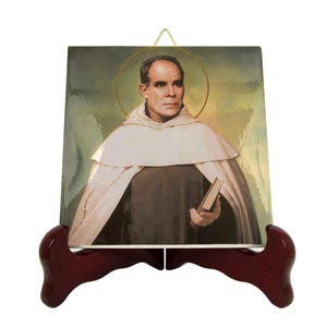 May include: A ceramic tile with a picture of a man in a brown robe and a white cowl holding a book. The tile is on a wooden stand.