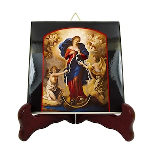 May include: A religious icon featuring a painting of a woman in red and blue robes, surrounded by angels. The image is set against a black background and displayed on a dark wooden easel. The painting depicts a religious scene.