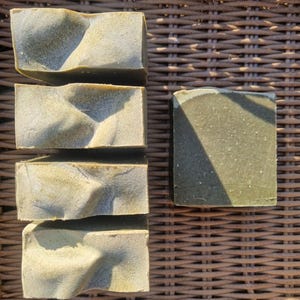 May include: Five rectangular bars of handmade soap with a textured, wavy surface and a light green colour. One additional bar is also present, with a flat top and a darker green hue. The soaps are arranged on a woven wicker surface.