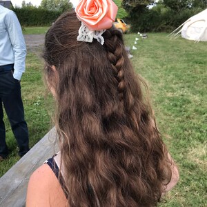 May include: A woman with long, brown, wavy hair wearing a peach-colored flower hair clip with white lace. The flower is made of satin and has a delicate, feminine design.
