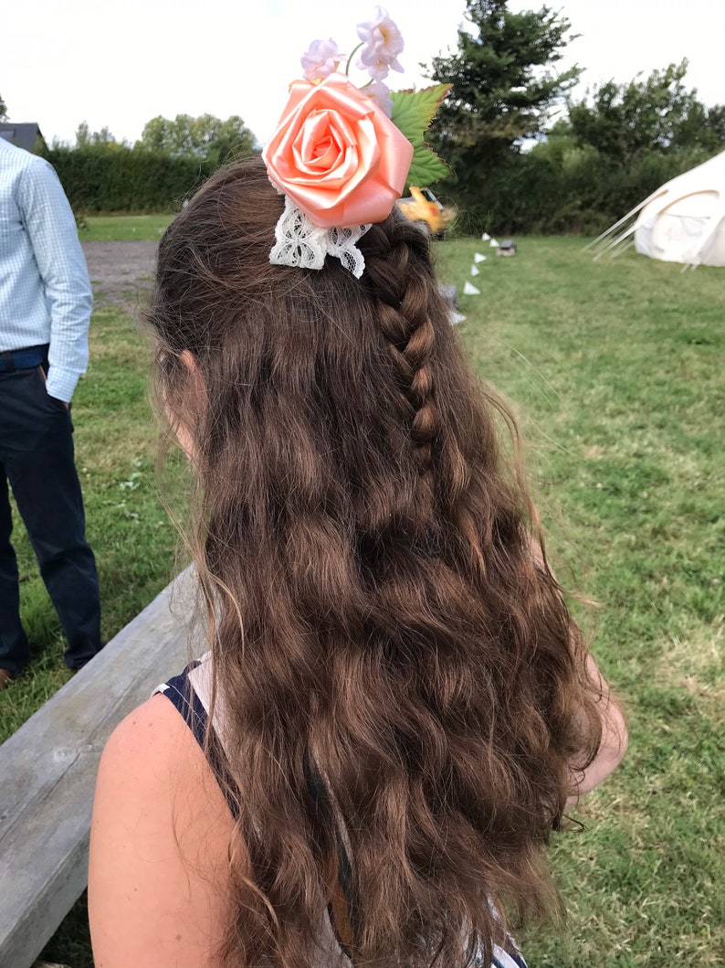 May include: A woman with long, brown, wavy hair wearing a peach-colored flower hair clip with white lace. The flower is made of satin and has a delicate, feminine design.