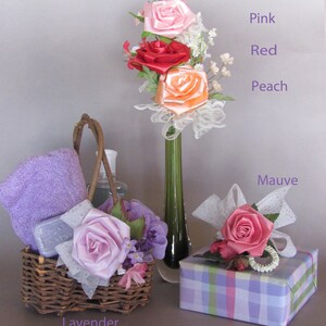May include: A gift basket with a purple towel, soap, and a lavender-colored satin rose. The basket is next to a vase with pink, red, and peach satin roses. A gift box with a mauve satin rose is in the foreground.