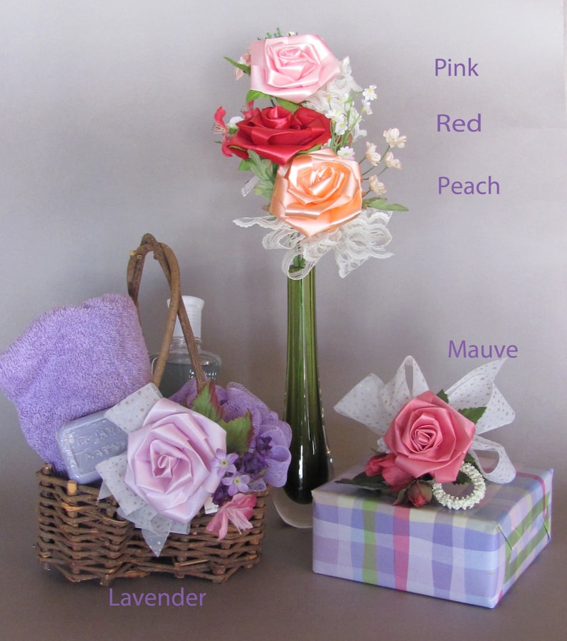 May include: A gift basket with a purple towel, soap, and a lavender-colored satin rose. The basket is next to a vase with pink, red, and peach satin roses. A gift box with a mauve satin rose is in the foreground.