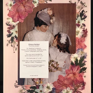 May include: A pink floral pressed flower frame with a photo of two women in white dresses. The frame is 11 inches by 14 inches and is designed for wedding invitations, photos, announcements, greeting cards, and custom calligraphy. The text "Picture Perfect Mounting System" and "Victoria" are visible in the image.